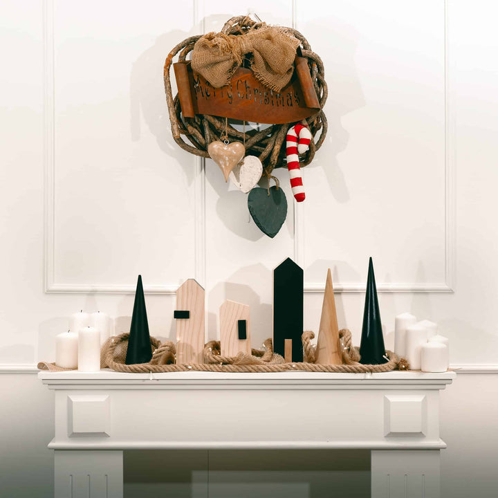 Decorative wall hanging with wooden houses and green cones on a white surface.