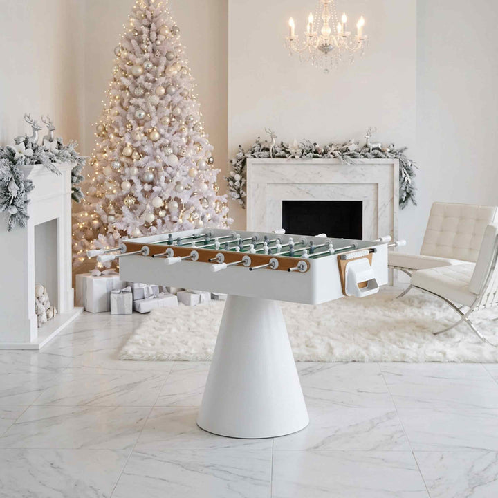 A white foosball table with a conical base and glass top, styled in a bright, modern holiday living room next to a decorated Christmas tree and fireplace.