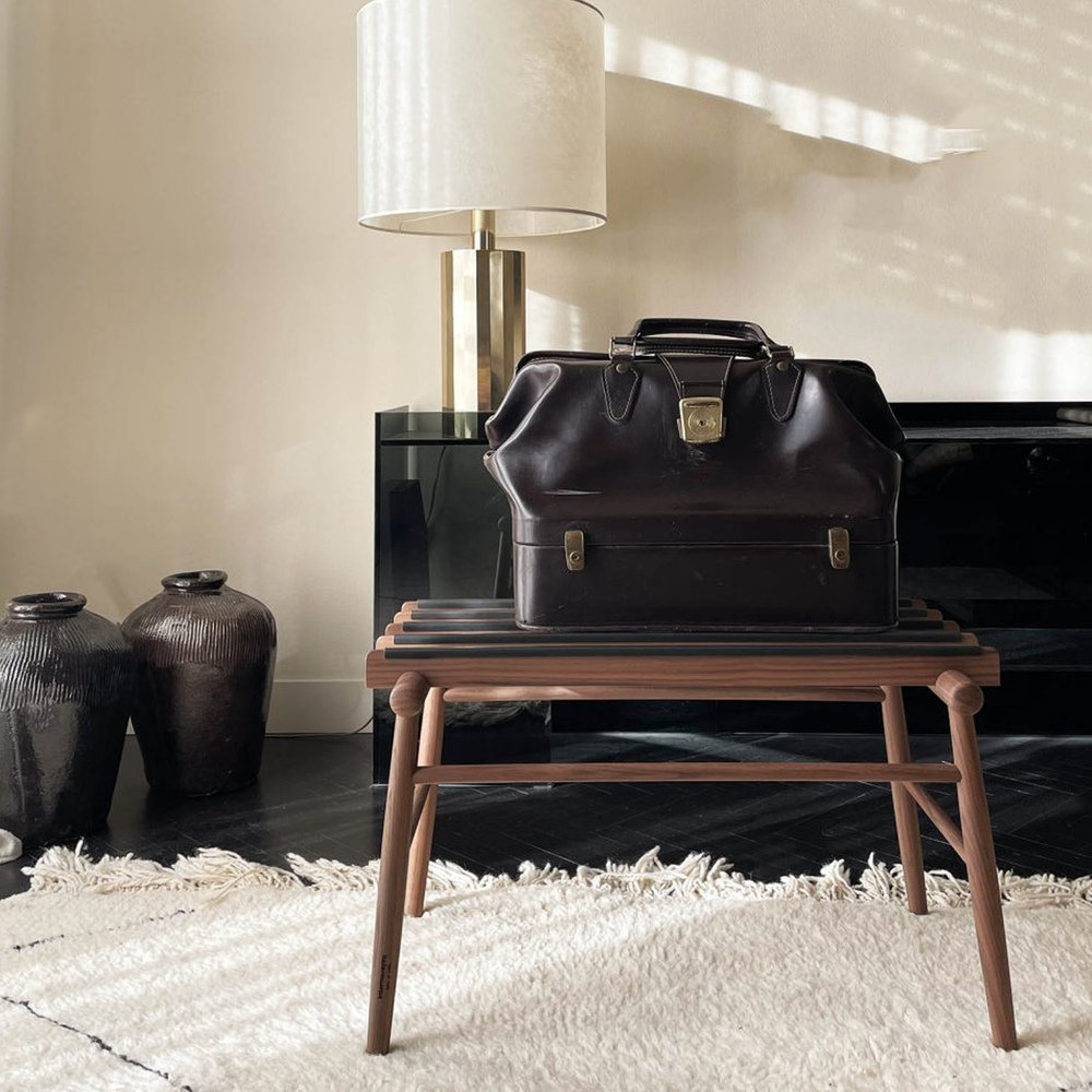 A dark brown leather travel briefcase with gold-tone hardware sits on a wooden bench in a styled interior setting with a lamp, vases, and a rug.