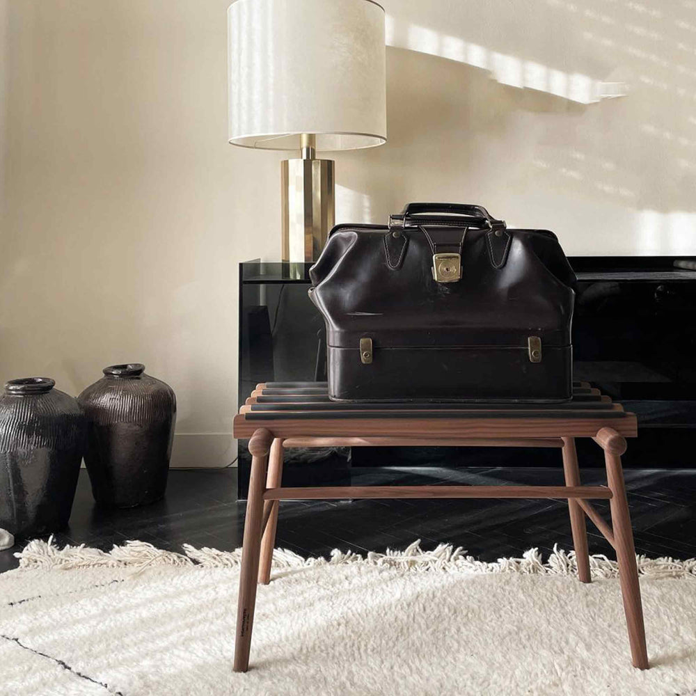 A dark brown leather travel briefcase with gold-tone hardware sits on a wooden bench in a styled interior setting with a lamp, vases, and a rug.