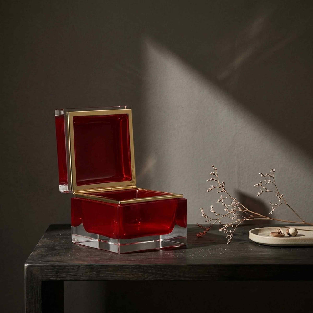 Open red glass box by Alessandro Mandruzzato on a dark wooden table beside dried stems and a small jewellery tray