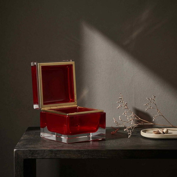 Open red glass box by Alessandro Mandruzzato on a dark wooden table beside dried stems and a small jewellery tray