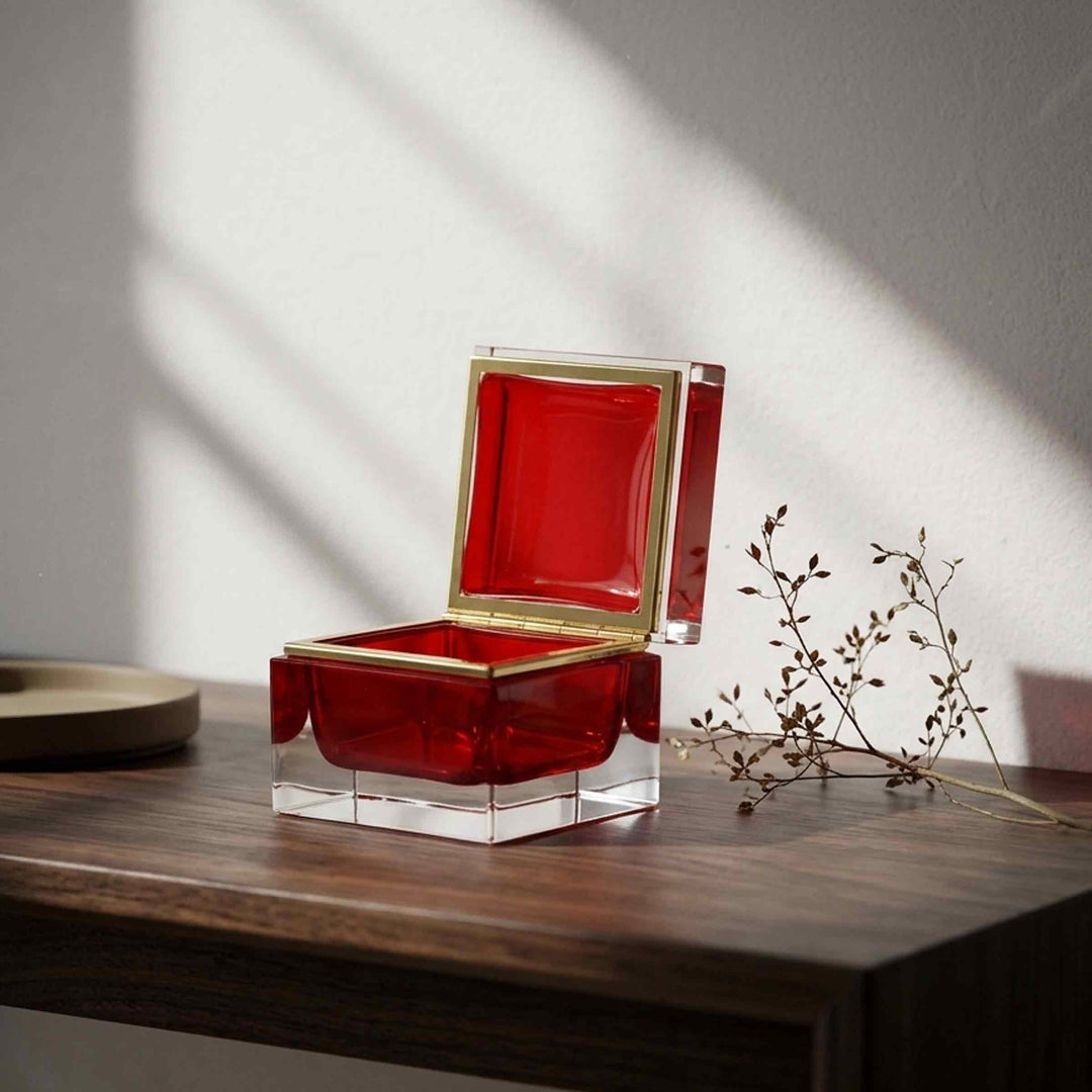 Open red square glass box by Alessandro Mandruzzato with gold-tone trim on a sunlit wooden table beside dried branches