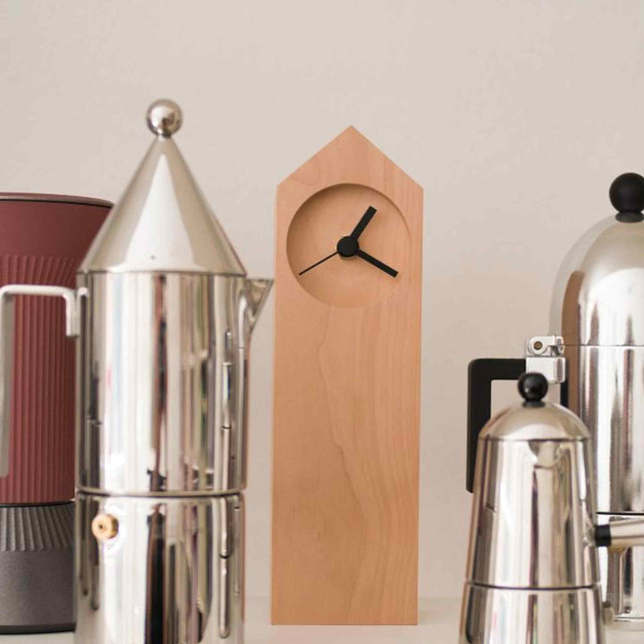 Osio table clock by Internoitaliano with a tall, house-shaped wooden body and black hands, displayed among metal coffee makers.