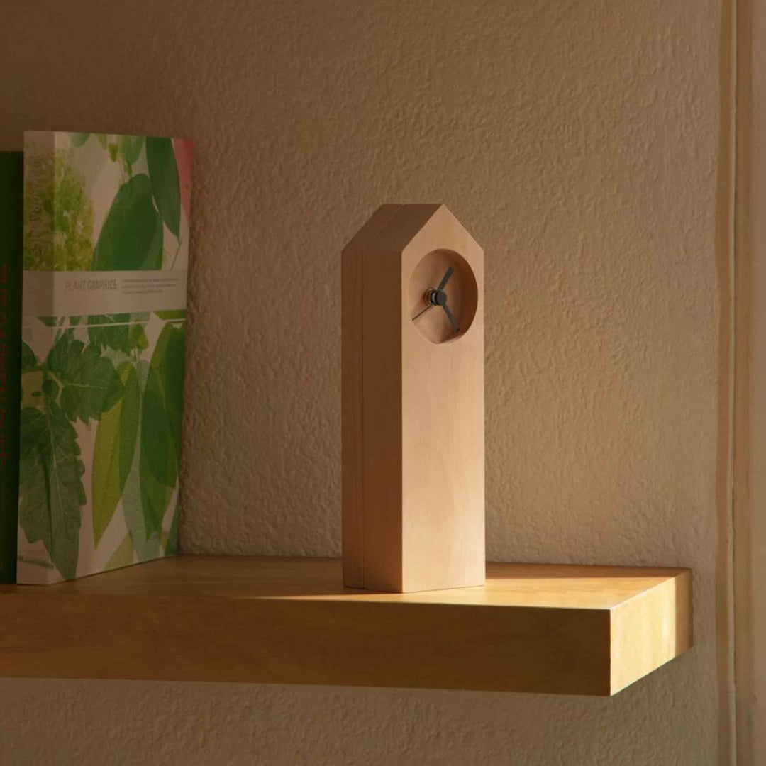 Osio house-shaped wooden table clock by Internoitaliano on a sunlit wooden shelf beside books.
