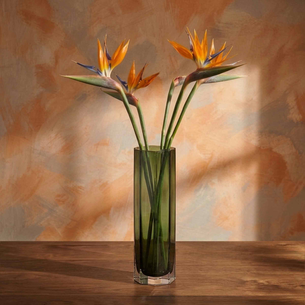 Tall green hexagonal glass vase by Alessandro Mandruzzato holding orange bird-of-paradise flowers on a wooden table.